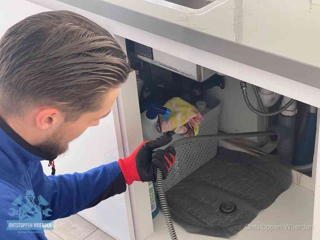 Een monteur voert een flexibele ontstoppingsveer in onder het keukenkastje, langs de sifon, om de gootsteen te ontstoppen in Woerden