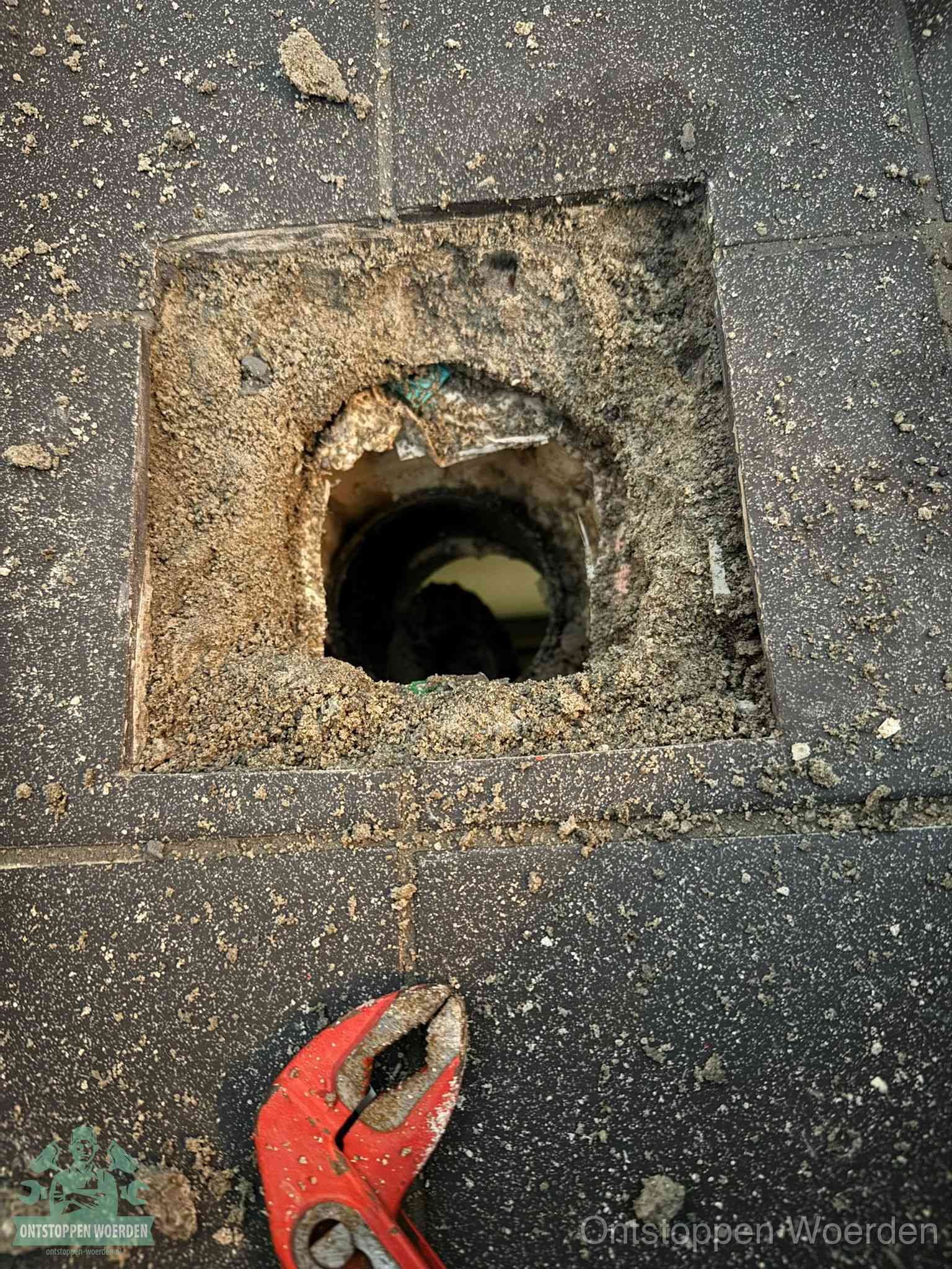 Een geopend rioolputje in een betontegelvloer met losgemaakt rooster en een zichtbare ronde afvoerbuis, het achterhalen waar de afvoerverstopping zit in Woerden