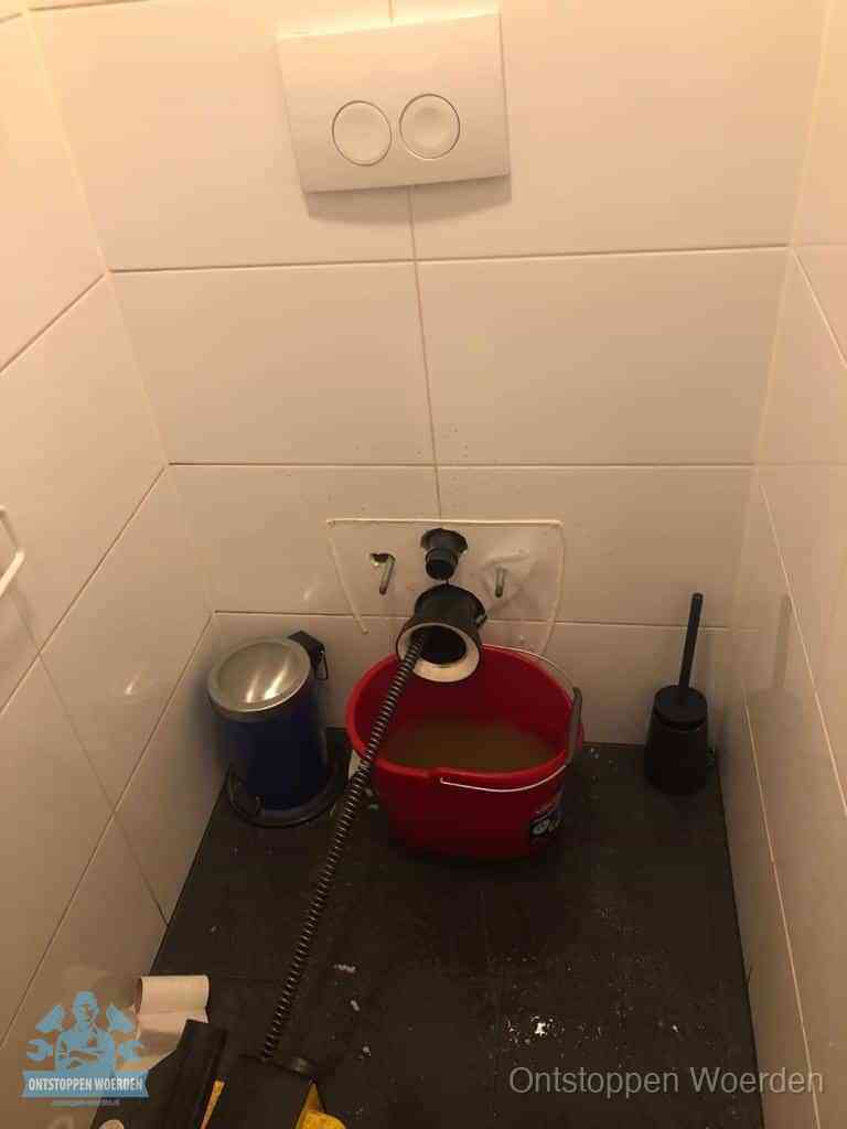 Een ontstopte wand-wc met verwijderde toiletpot waar een ontstoppingsveer in de wanduitgang verdwijnt, het achterhalen waar de verstopping zit in Woerden
